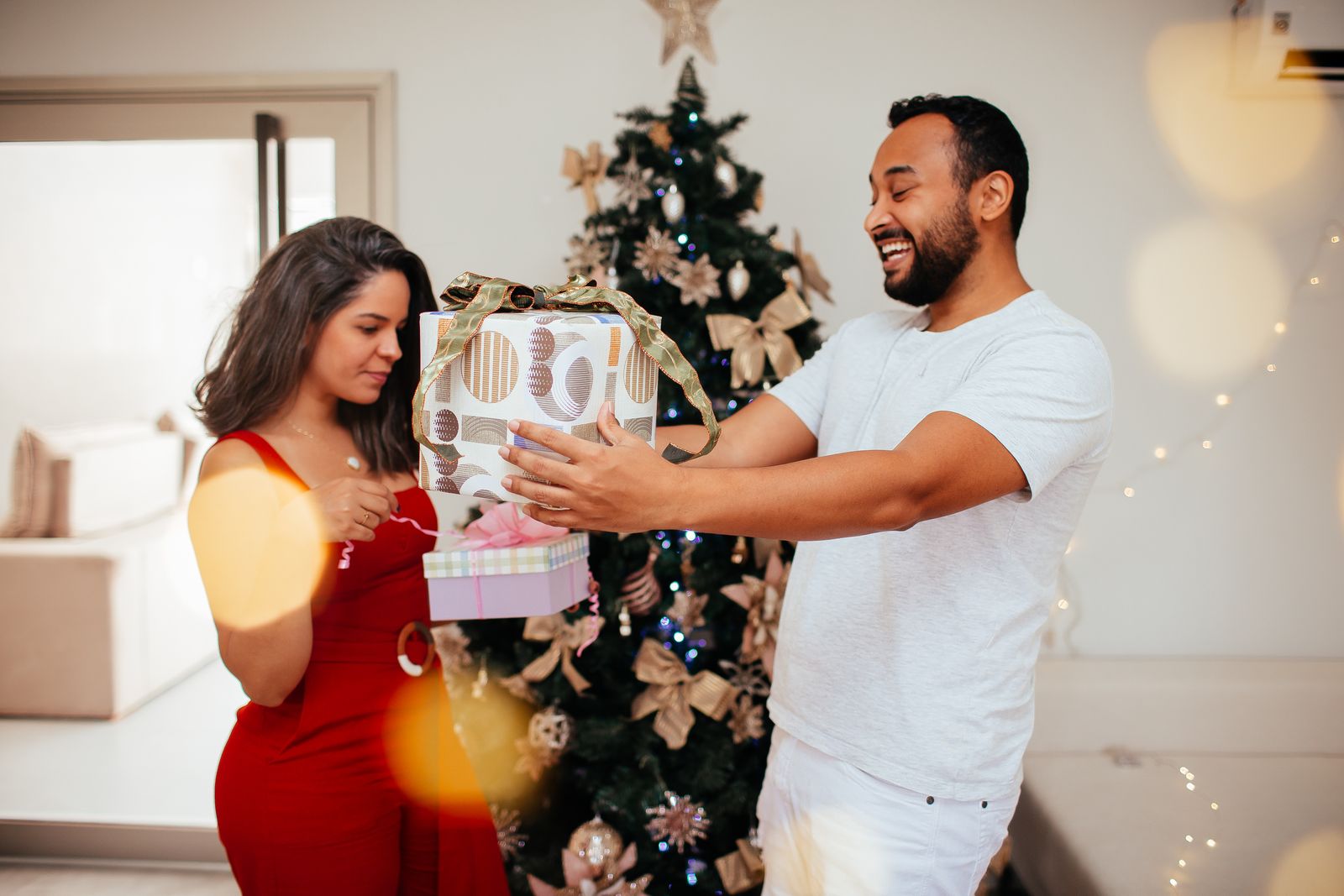 A couple laughing as they exchange gifts in front of a Christmas tree