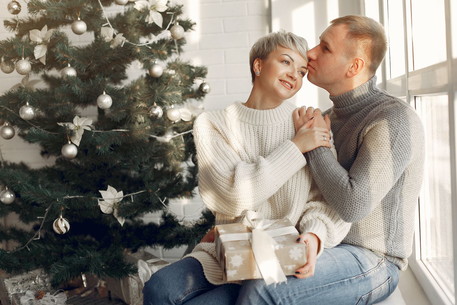 A couple in sweaters sitting close together by a window next to a Christmas tree