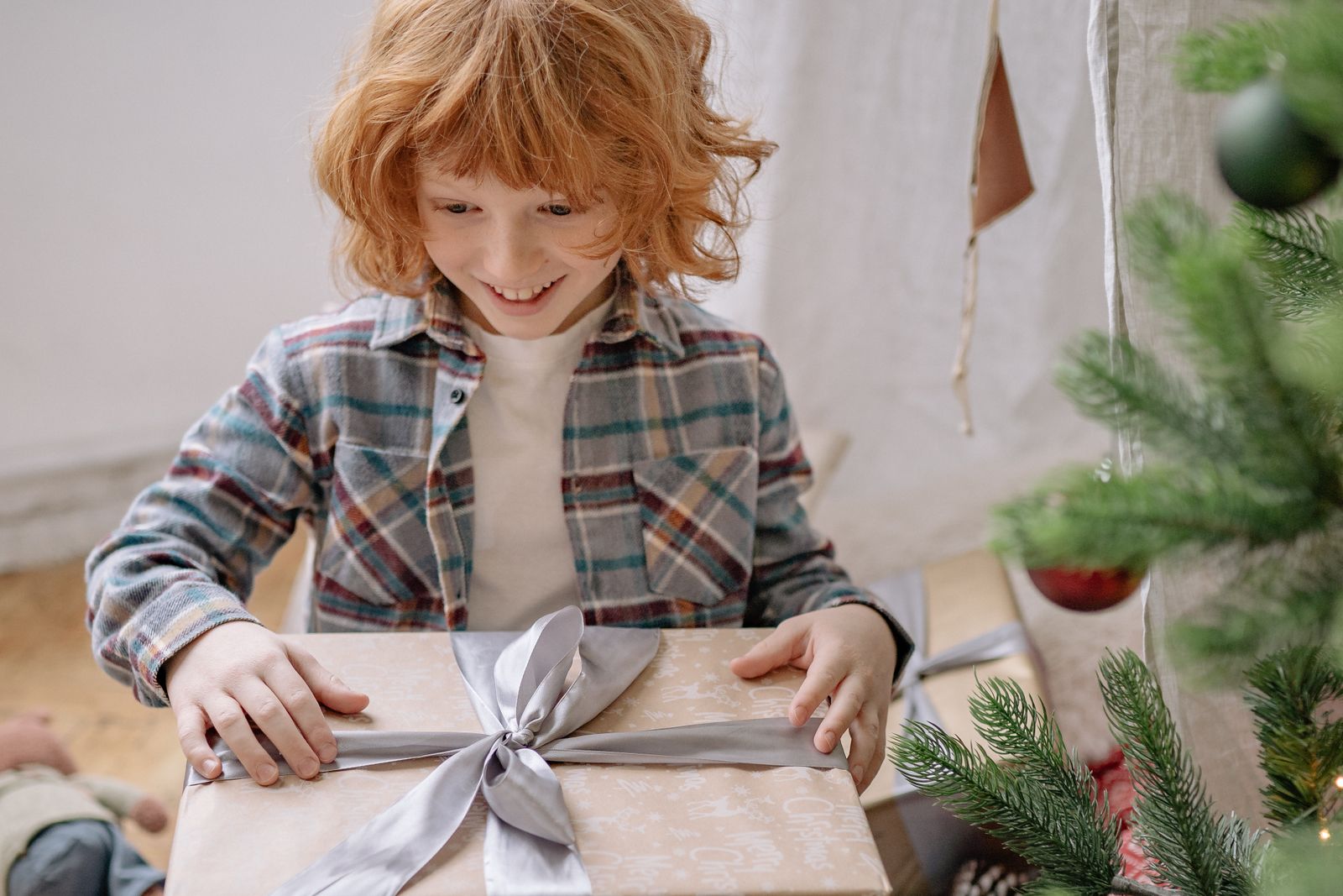 A smiling child with curly red hair opening a wrapped gift tied with a silver ribbon next to a decorated Christmas tree