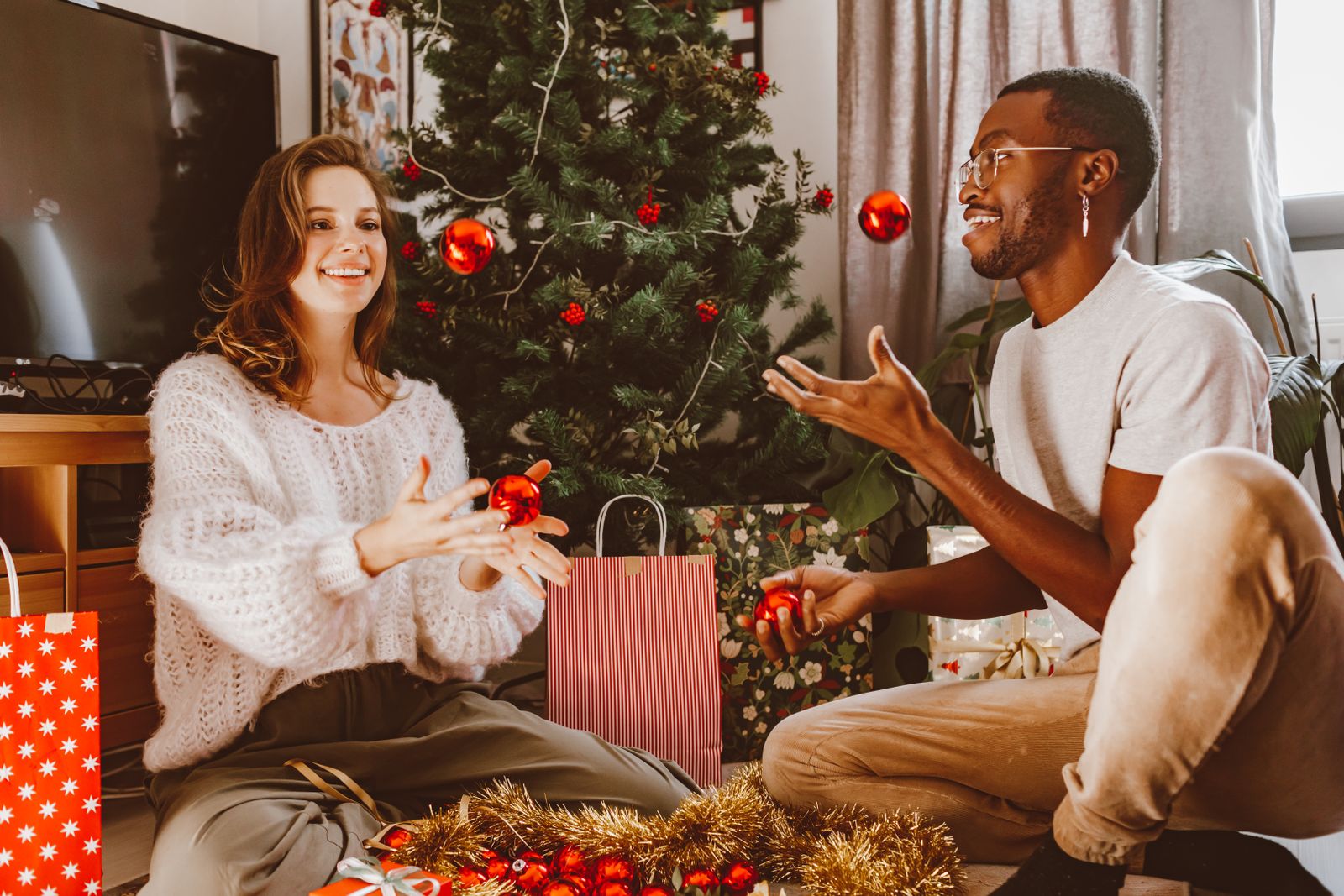 A smiling couple tossing red ornaments to each other beside a decorated Christmas tree