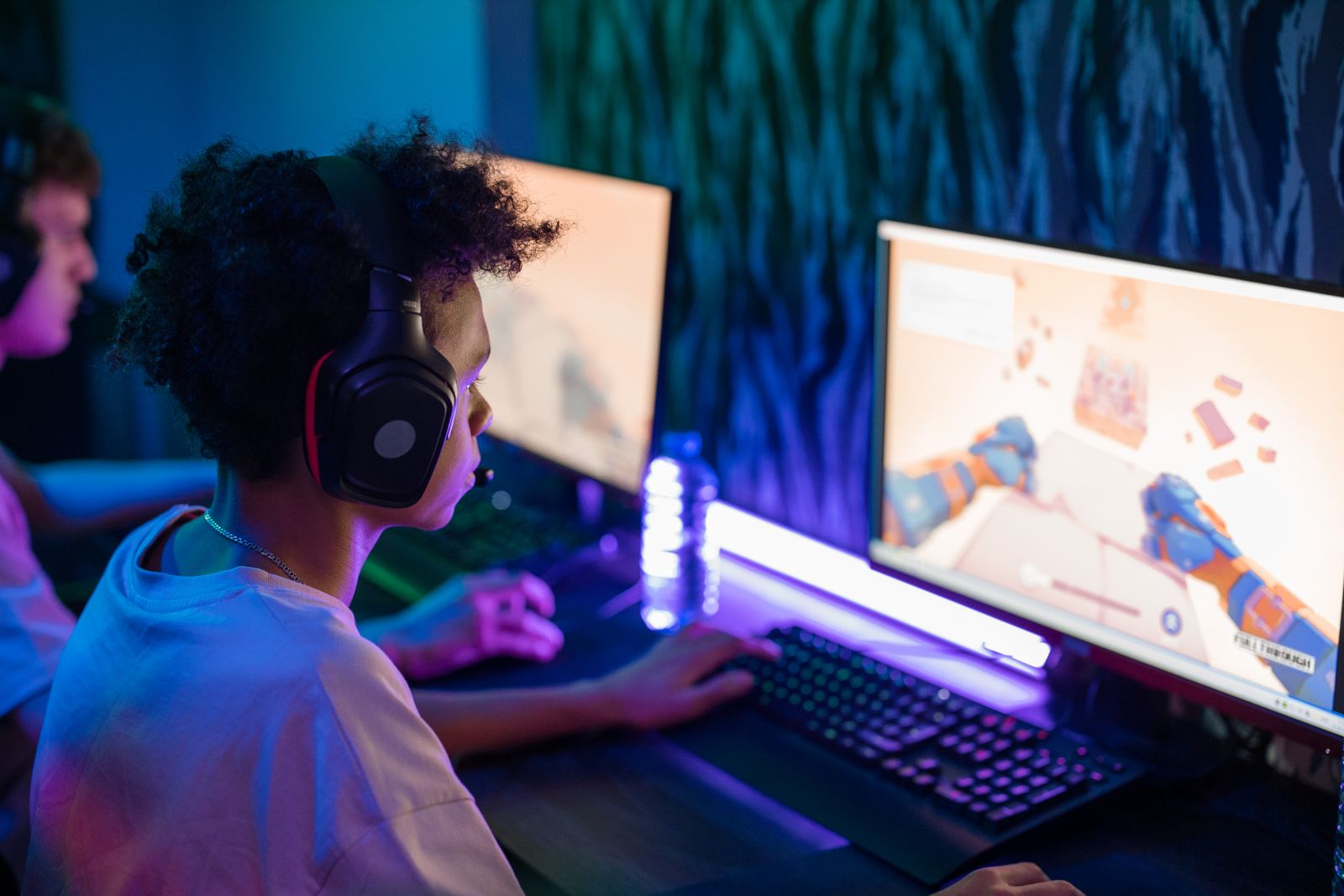 A teenage boy wearing headphones and playing a game on a computer in a dim room lit with blue and purple light