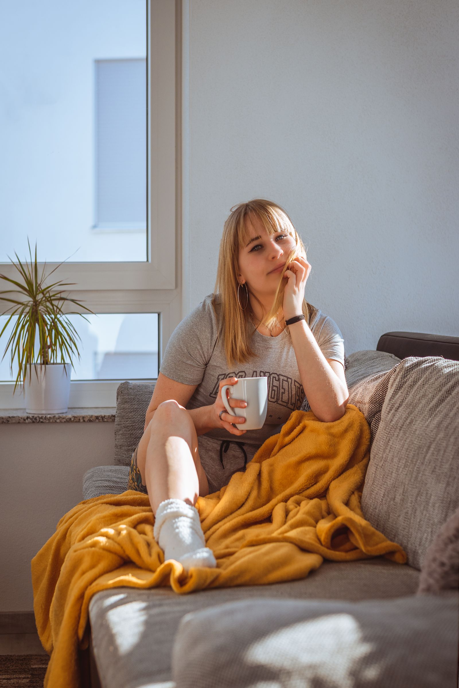 A woman in sunlight curled up on a grey sofa under a yellow blanket, holding a white mug